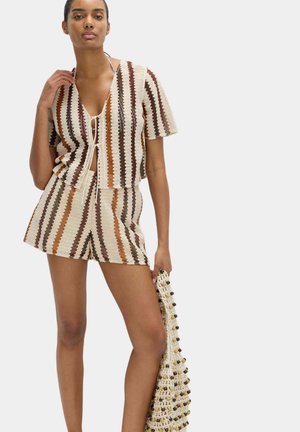 Kobieta ubrana w prążkowany krótki rękaw swetrowy i pasujące szorty, trzymająca torebkę z dzianiny z koralikami na jednolitym tle.