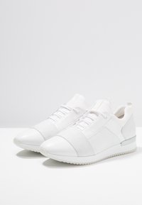 Paire de baskets basses blanches avec bande élastique sur le devant, fermeture à lacets, et semelles en caoutchouc texturées sur fond gris uni.