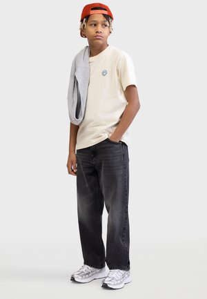 Έφηβος με μπεζ μπλουζάκι με λογότυπο με χαμογελαστό πρόσωπο, μαύρα τζιν, ασημένια αθλητικά παπούτσια, πορτοκαλί καπέλο φοράει ανάποδα, γκρι φούτερ πάνω στον ώμο, κοιτάζει αριστερά.