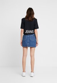 Kvinna som står med ryggen mot, iklädd en svart croppad T-shirt med texten "JEANS" och en blå denim mini kjol tillsammans med vita sneakers.