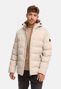 Doudoune beige avec capuche, présentant un design matelassé, des poches latérales zippées et un intérieur en polaire, portée par-dessus un pull beige.