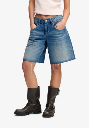 Person iført falmede denim knælange shorts med flossede kanter, en hvid cropped top og mørkebrune støvler med spænde.