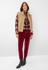 Beige Sherpa-Weste über einer gemusterten bordeauxfarbenen Bluse, kombiniert mit burgunderfarbenen Cordhosen und schwarzen Ankle Boots. Sanfte Texturen und ergänzende Farben.