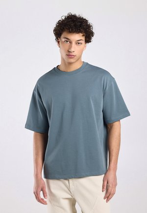 Young man with curly hair wearing a loose-fitting blue-gray t-shirt and light beige pants, standing against a plain light background.