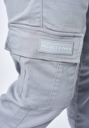 Pantaloni cargo grigi con una texture liscia, dotati di una tasca laterale con etichetta "PROJECT X PARIS". Le cuciture visibili ne esaltano il design.