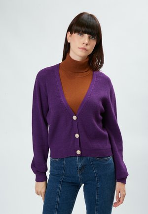 Cardigan lavorato a maglia di un vivace colore viola con scollo a V profondo, caratterizzato da bottoni simili a perle e maniche lunghe, indossato sopra un dolcevita marrone.