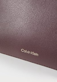 Suprafață din piele maronie cu un logo auriu "Calvin Klein" evidențiat prin embosare, subliniind brandul și textura.
