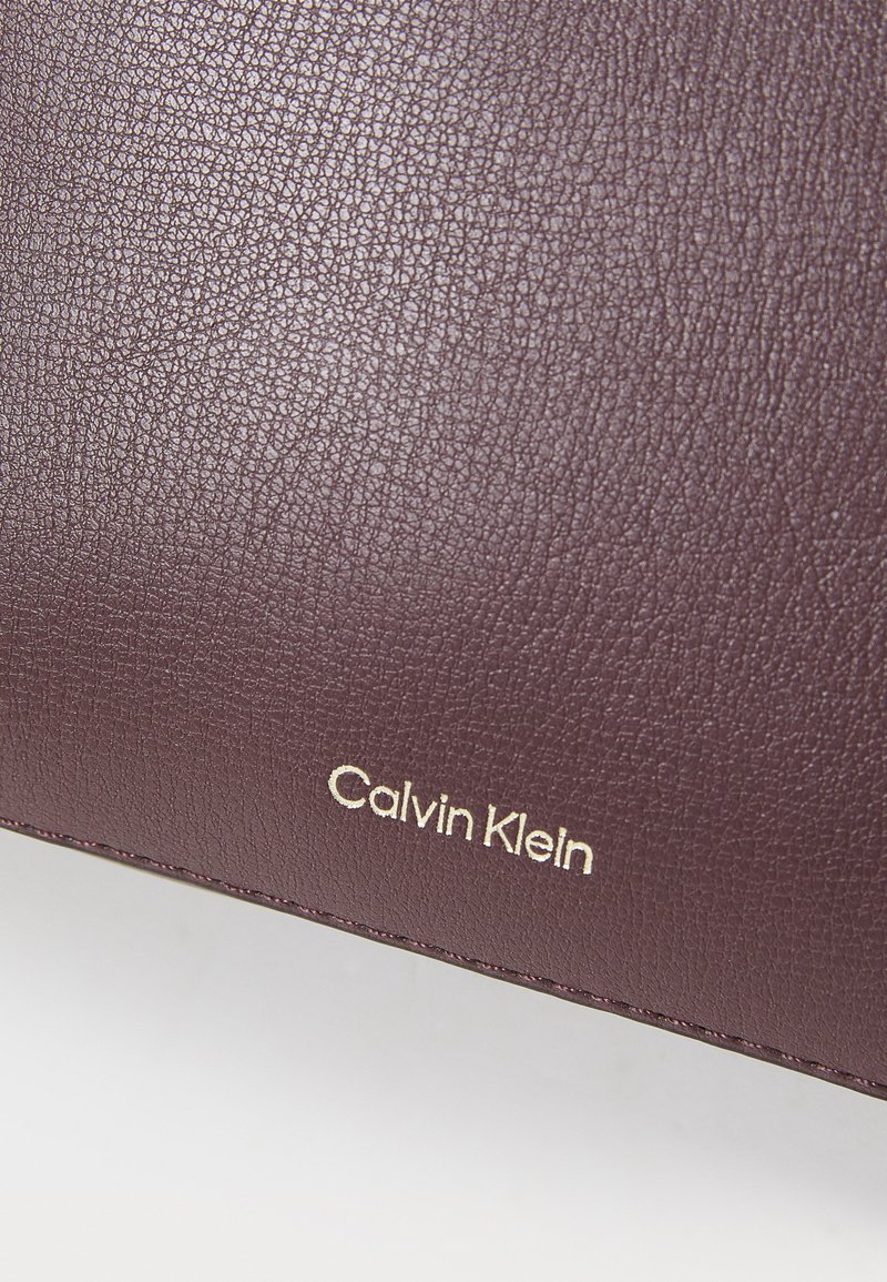 Superficie in pelle color bordeaux con un prominente logo dorato "Calvin Klein" impresso, che mette in evidenza il branding e la texture.