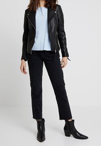 Černá kožená motocyklová bunda přes světle modrý svetr, doplněná černými zkrácenými džíny a botami s špičatou špičkou za kotníky. Hladké textury a moderní silueta.