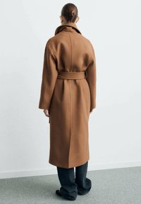 Bruine wollen trenchcoat met een ceintuur in de taille, grote kraag en lange mouwen, gecombineerd met donkere spijkerbroek en zwarte schoenen.