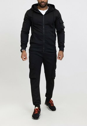 Haut à zip noir avec capuche, poches avant et tissu texturé, associé à un pantalon cargo noir avec poches latérales, porté avec des baskets noires et orange.