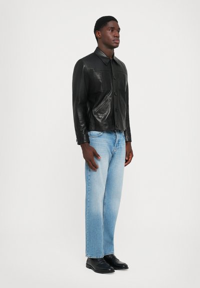 Mand står iført en sort læderjakke med knapper, lyseblå jeans og sorte lædersko mod en almindelig hvid baggrund.