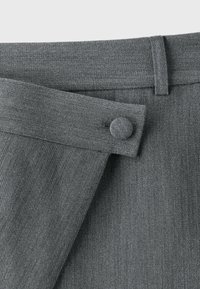 Taille d’un pantalon en tissu gris avec un rabat sécurisé par un bouton recouvert de tissu et une boucle de ceinture visible sur le côté droit.