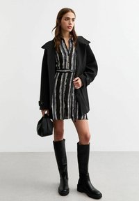 Zwarte wollen jas over een gestreepte jurk in blauw, wit en zwart. Tot de knie reikwijdte zwarte leren laarzen. Model houdt een kleine zwarte tas vast.