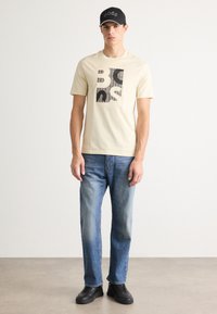 Man wearing beige graphic-print short-sleeve t-shirt, blue jeans, black sneakers, and black cap with text, standing on a plain floor against white wall.