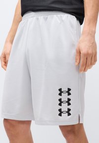 Κοντινή λήψη ατόμου που φοράει αθλητικά σορτς ανοιχτού γκρι χρώματος με τρία στοιβαγμένα μαύρα λογότυπα Under Armour στον αριστερό μηρό και μαύρο διχτυωτό μπλουζάκι.