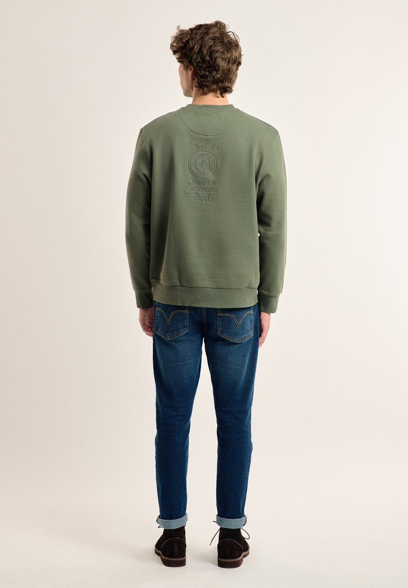 Sudadera verde oliva con textura suave, que presenta un detalle de logo bordado en la parte trasera. Combinada con jeans de mezclilla azul y zapatos oscuros.