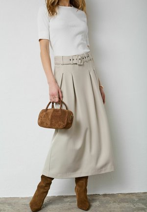 Femme portant un haut blanc à manches courtes, une jupe plissée beige taille haute avec une large ceinture, des bottes en daim marron, tenant un petit sac à main en daim marron.