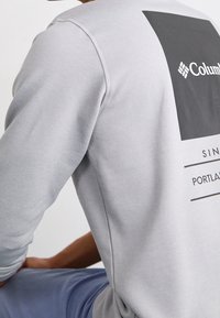 Persona vestida con un sudadera gris claro de Columbia con un logo cuadrado negro y texto, sentada con un brazo apoyado en la rodilla doblada.