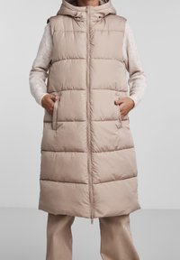 Béžová prošívaná vesta s kapucí, má zipový přední díl, postranní kapsy a horizontálně prošívané části pro větší teplo a texturu.