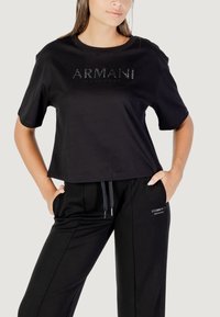 Camiseta de algodón negra con mangas cortas, que presenta un logo brillante de "ARMANI EXCHANGE" en el frente. Combinada con pantalones de chándal negros.