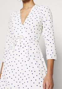 Robe blanche à pois bleus, avec un décolleté en V, des manches trois-quarts et un détail de taille nouée. Tissu lisse et léger.
