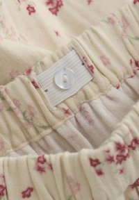 Stoff mit Blumenmuster in Cremefarbe und kleinen roten Blumen. Verfügt über einen weichen, gerafften Bund und einen Kunststoffknopf zum Verschließen.