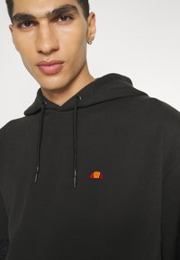 Sudadera negra con cordones, con un pequeño logo bordado en naranja y rojo. Tejido suave, corte relajado y diseño minimalista.
