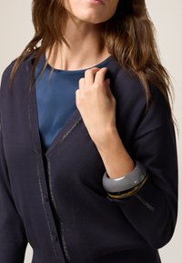 Cardigan blu navy con dettagli brillanti, indossato sopra una maglietta blu. La modella indossa un bracciale spesso grigio e nero al polso.