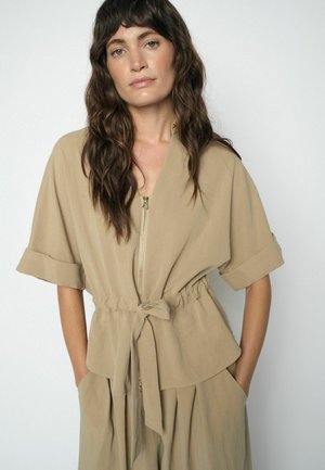 Woman with long wavy hair wearing a beige short-sleeve zip-up top with a tied waist and matching wide-leg pants, standing with hands in pockets.