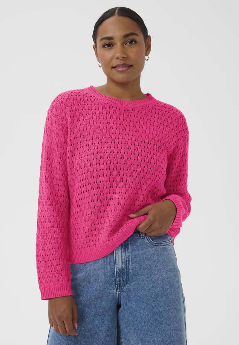 Femme portant un pull en maille texturée rose vif et un jean bleu, debout devant un fond clair uni.