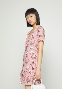 Vestido floral rosa com mangas curtas, decote em V profundo, com bainha em babados e padrões de rosas. Bolsa branca segurada na mão direita.
