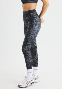 Hoge taille leggings in een zwart-grijs luipaardpatroon, gemaakt van rekbaar materiaal, slim fit ontwerp, gecombineerd met witte sokken en sneakers.