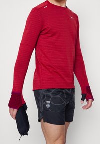 Personne portant un haut de sport rouge à manches longues avec des trous pour les pouces et un short noir à motifs, tenant une pochette noire dans une main.