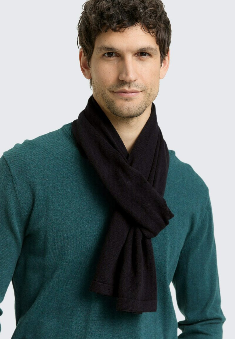 Sciarpa nera lavorata a maglia avvolta intorno al collo di una persona, abbinata a un maglione verde, che mostra una texture liscia e un design semplice.