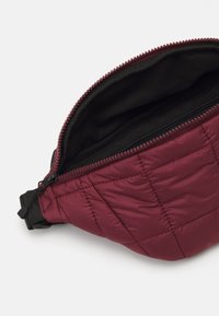 Burgundy quilted waist bag with a zipper and black interior lining; features durable fabric and adjustable strap for secure fit.