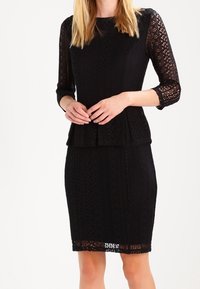 Sort blondekjole med trekvartærmer, peplum talje og tætsiddende nederdel. Har indviklede mønstre og en tekstureret finish.