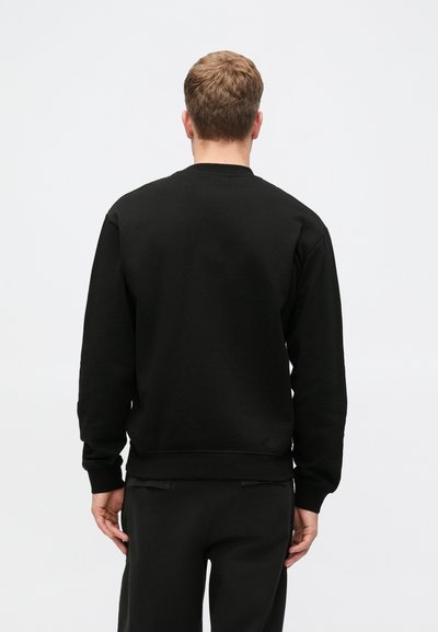 Hombre con cabello corto castaño de espaldas, vistiendo un sudadera negra lisa y pantalones negros, frente a un fondo blanco.