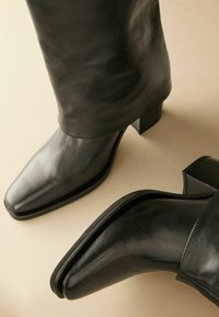Botines de cuero negro hasta el tobillo con punta cuadrada, tacón alto y textura suave. Presenta un cañón ajustado y mínimos acentos.