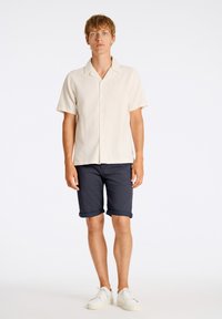 Camisa de manga corta color beige con botones en la parte frontal, combinada con pantalones cortos azul marino con dobladillo rollado. El modelo lleva zapatillas blancas.