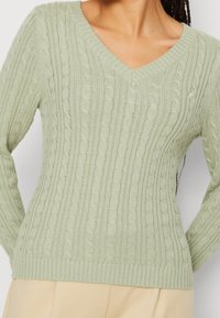 Hellgrüner Kabelstrickpullover mit V-Ausschnitt, langen Ärmeln, geripptem Saum und Manschetten. Verfügt über strukturierte Muster im gesamten Stoff.