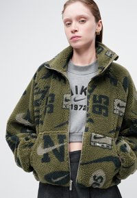 Roheline fliisk jacket, millel on lukuga esitaskud, suured taskud ja tumedamate varjunditega logo muster. Peal on lühikeste varrukatega hall sweatshirti.