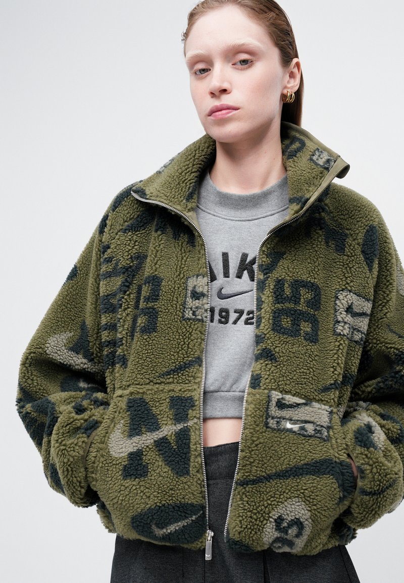 Grüne Fleecejacke mit Reißverschluss, großen Taschen und einem Muster aus Logos in dunkleren Farbtönen. Getragen über einem kurzen grauen Sweatshirt.