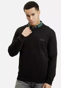 Czarna bluza z teksturowanej tkaniny, z okrągłym dekoltem i małym logo na piersi, uzupełniona zieloną koszulą w kratę noszoną pod spodem.