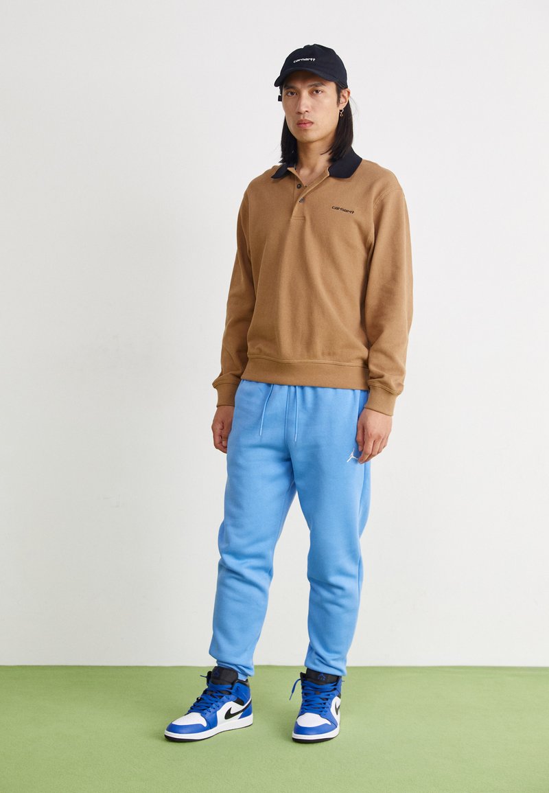 Brauner Pullover mit schwarzem Kragen, hellblaue Jogginghose und blau-weiße Sneaker. Schlichtes Design mit sichtbarem Markenlogo auf der Kleidung und Mütze.