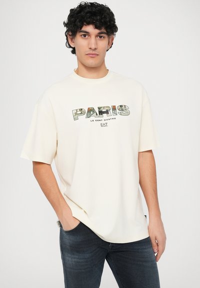 Krämfärgad oversized t-shirt med "PARIS" grafik i grönt och svart; kombinerad med mörka jeans. Avslappnad passform och mjuk textur.