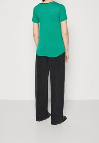 Camiseta verde de manga corta con dobladillo redondeado, de tela texturizada, combinada con unos jeans negros holgados y calzado negro, vista desde atrás.