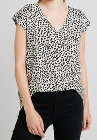 Donna che indossa un top senza maniche bianco a V con macchie nere irregolari e pantaloni neri a vita alta.