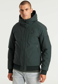 Chaqueta acolchada verde oscuro con capucha, presenta un cierre frontal con cremallera, bolsillos laterales y un bolsillo en el pecho. El material parece suave con una cintura texturizada.