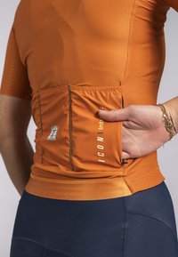 Oranje sporttop met korte mouwen, textuurstof en een achterzak. Benadrukt logo detail en platte naden. Gepaard met donkere leggings.